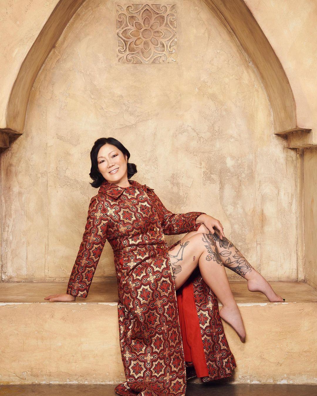 A photo showing Margaret Cho in an embroidered slit dress, sitting on a cemented surface.
