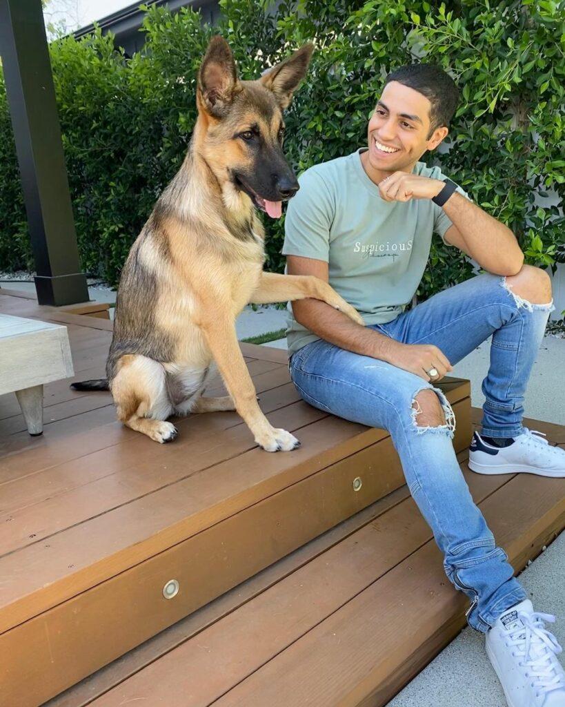 Mena Massoud sits beside his dog, who has his paw on his arm.