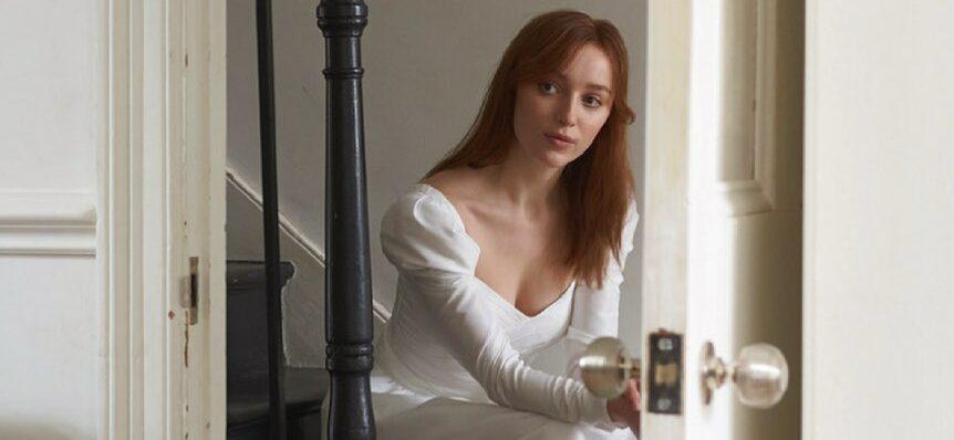 A photo showing Phoebe Dynevor in a white silk dress sitting on a sofa.