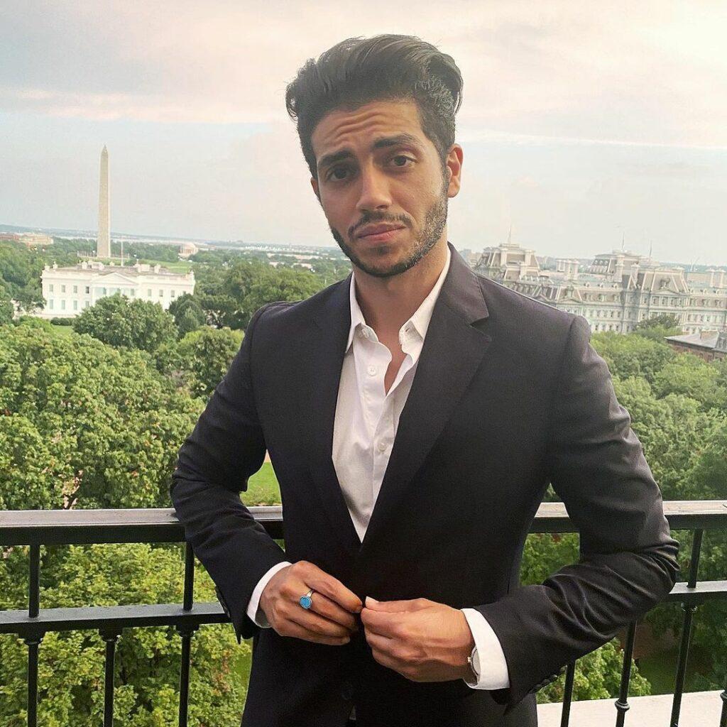 Mena Massoud poses for a photo on a balcony while buttoning a suit.