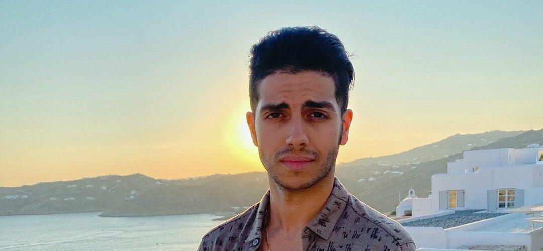 Mena Massoud poses in front of a sunset while in Greece.