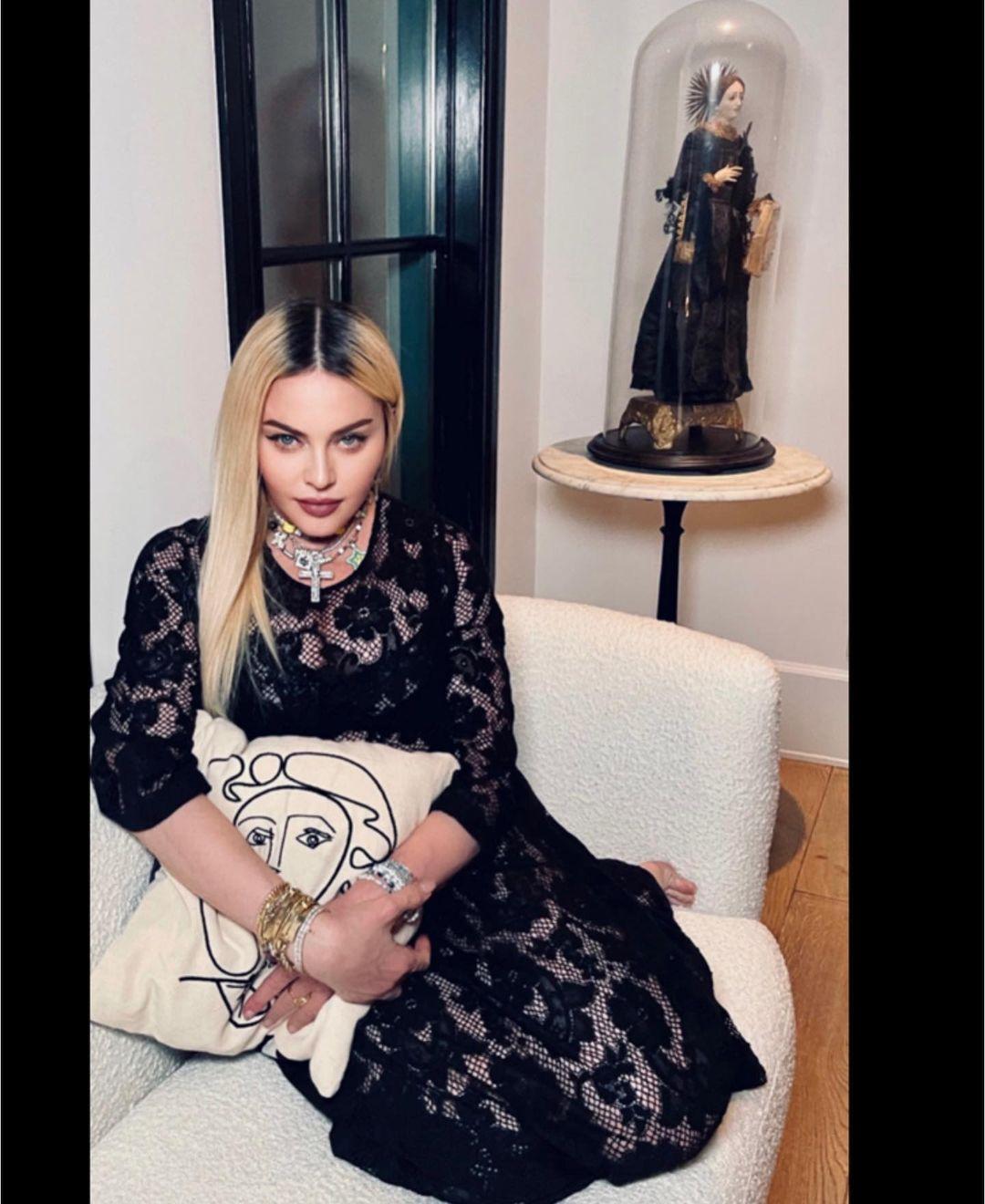 A photo of Madonna sitting on a sofa in a black lace dress.