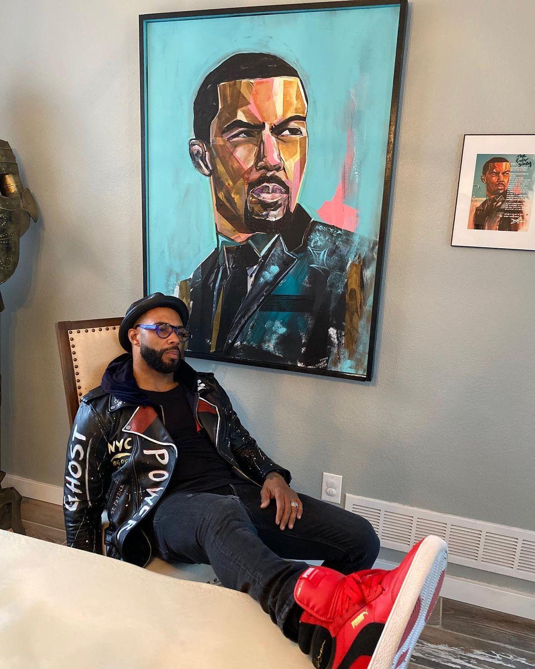A photo showing Omari Hardwick sitting beside a drawn portrait on him hanging on the wall.
