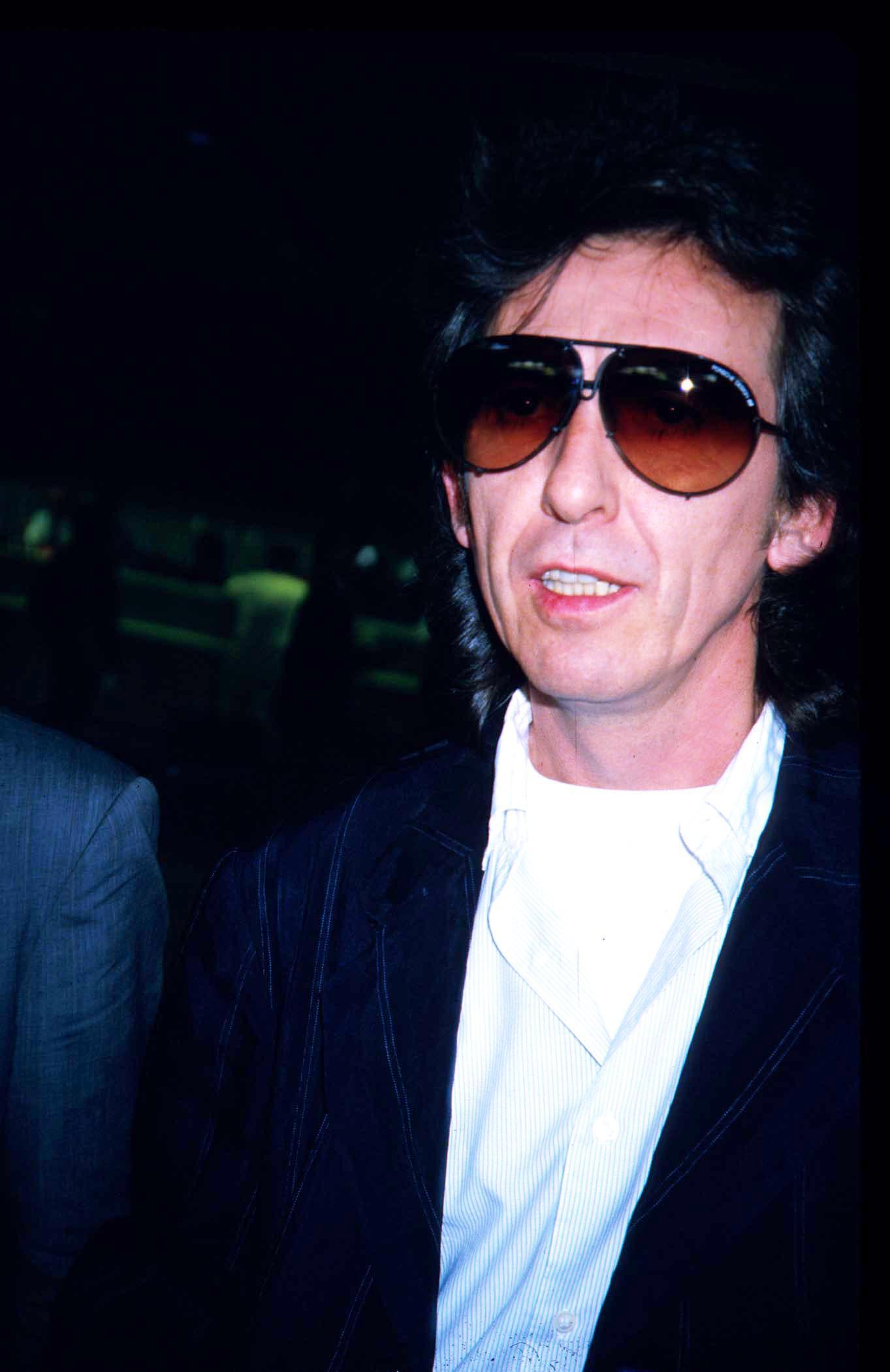 A photo showing George Harrison in a black suit, with a white inner T-shirt, and a pair of sunglasses.