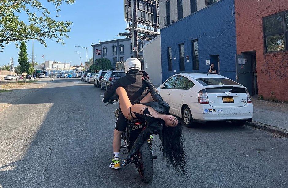 Lourdes Leon riding a motorcycle in a black bikini top, thong, leather jacket, and boots.