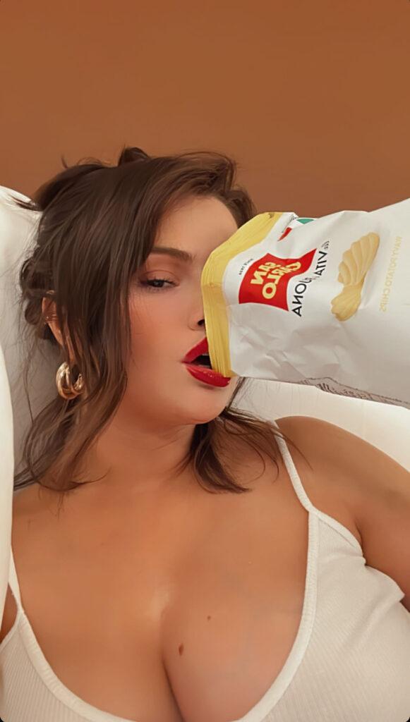 Selena Gomez eating a bag of chips in a low-cut white tank top.