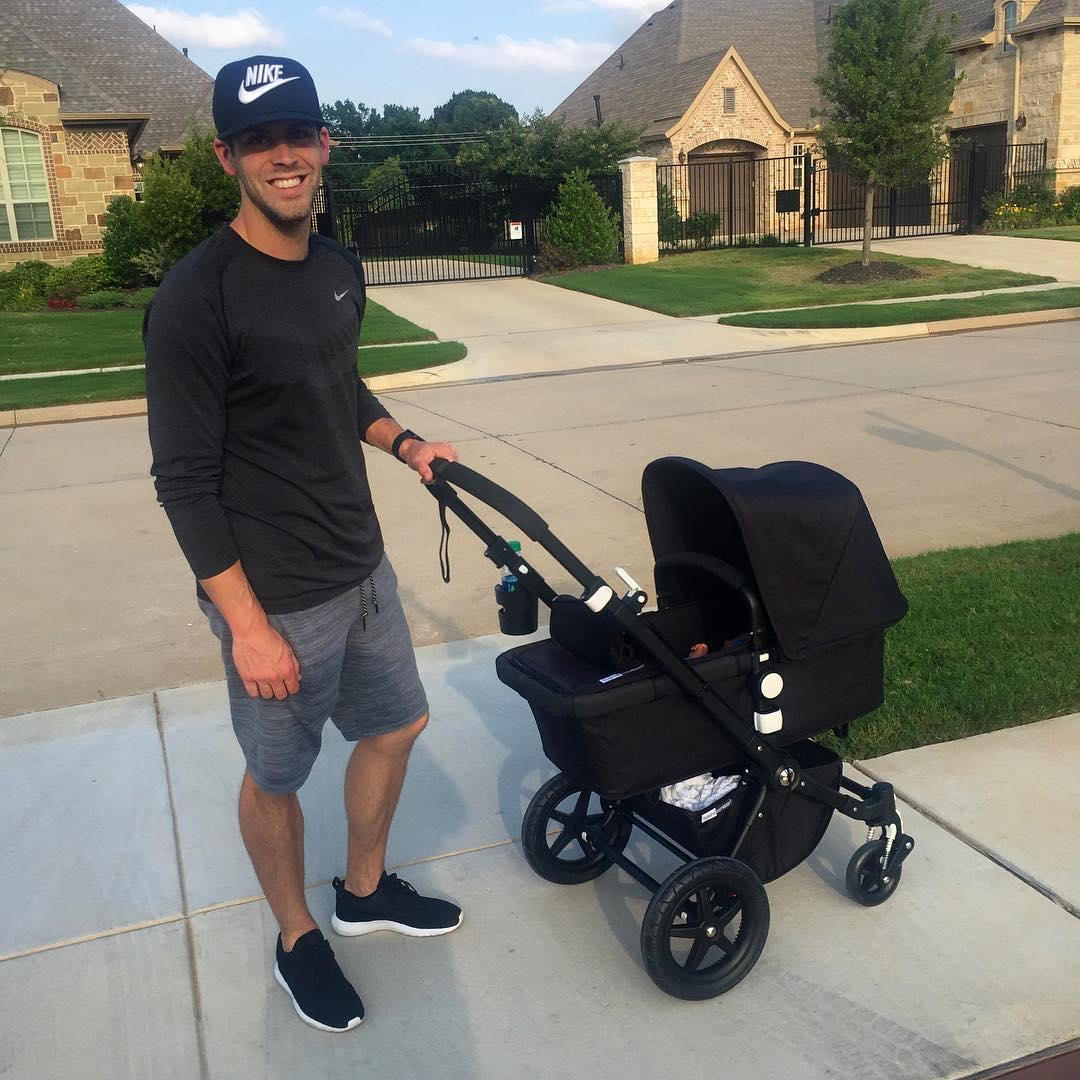 Justin Tucker wearing a black baseball hat, black shirt, and grey shorts, smiling while holding a baby stroller