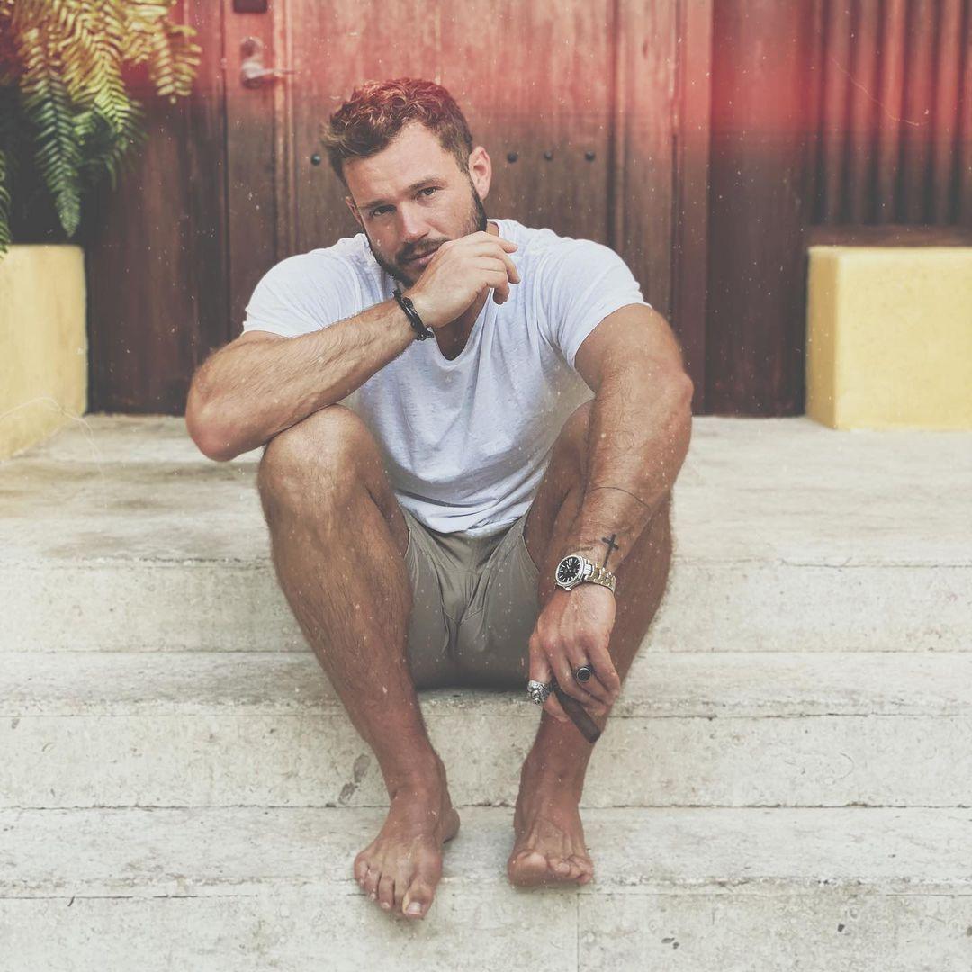 A photo showing Colton Underwood in a white shirt and brown short sitting on a short staircase