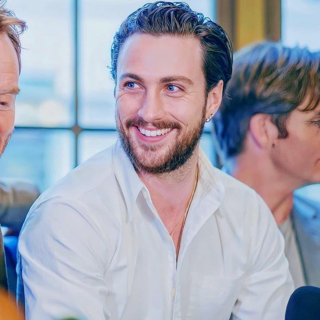 A photo of Aaron Taylor-Johnson smiling profusely at the camera in a white T-shirt