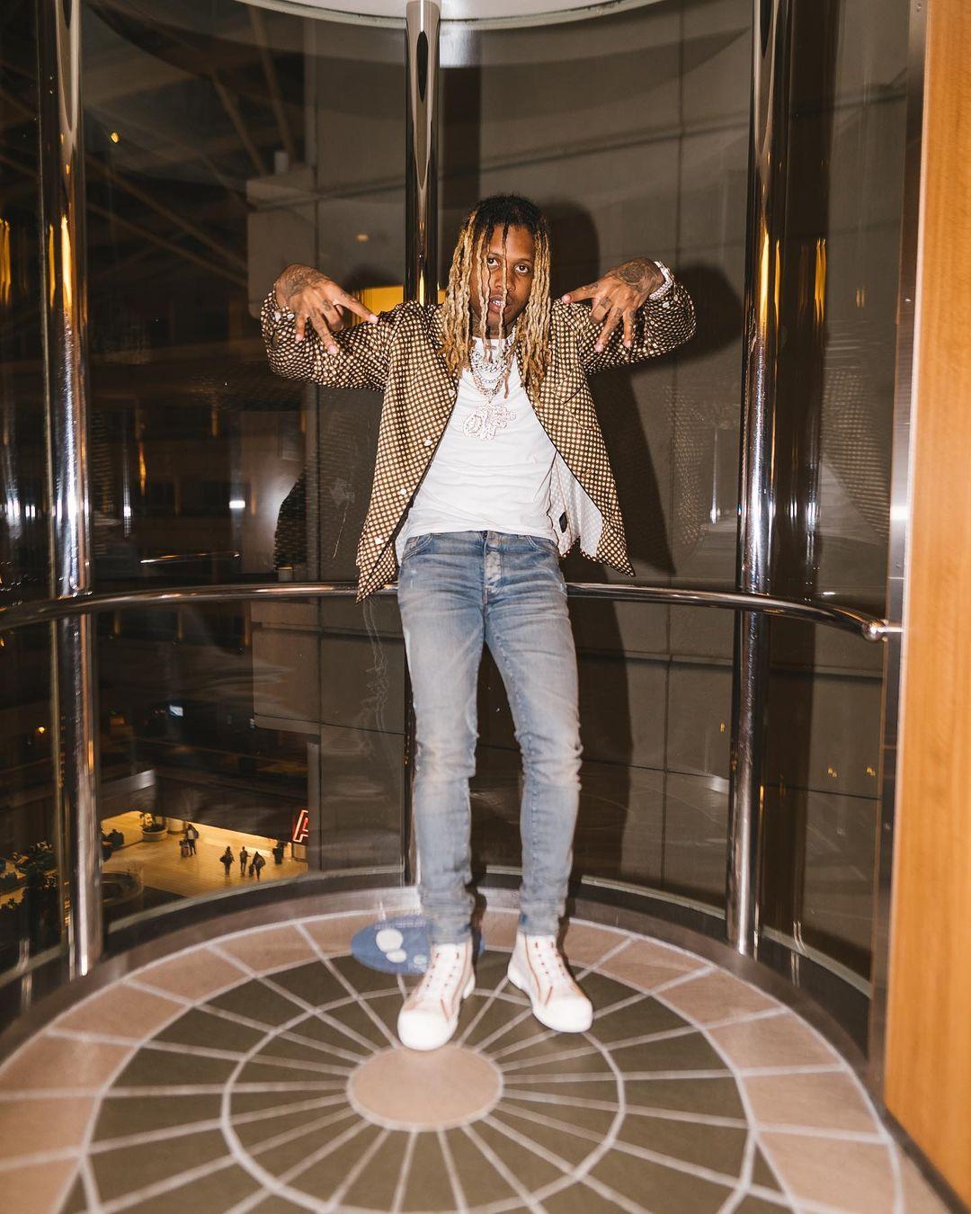 A photo showing Lil Durk in a brown jacket over a white round-neck and denim pant, with white sneakers to match.