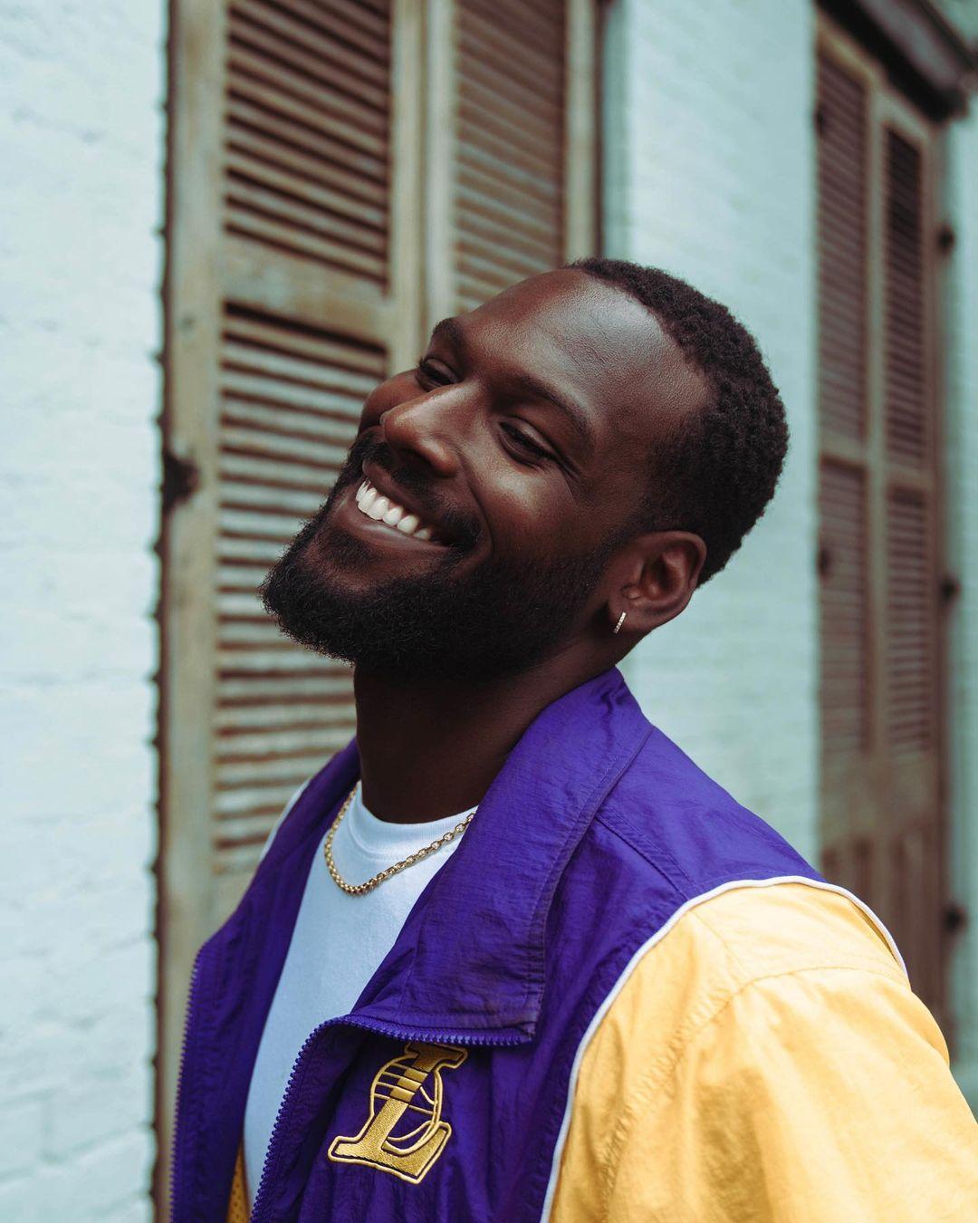 Kofi shows off a smile in this photo of him sporting a purple and yellow print sweater