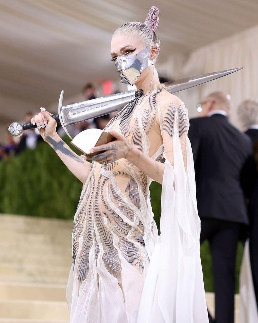 A photo showing Grimes dressed in a silver-themed dress with a shiny sword hanging over her shoulder