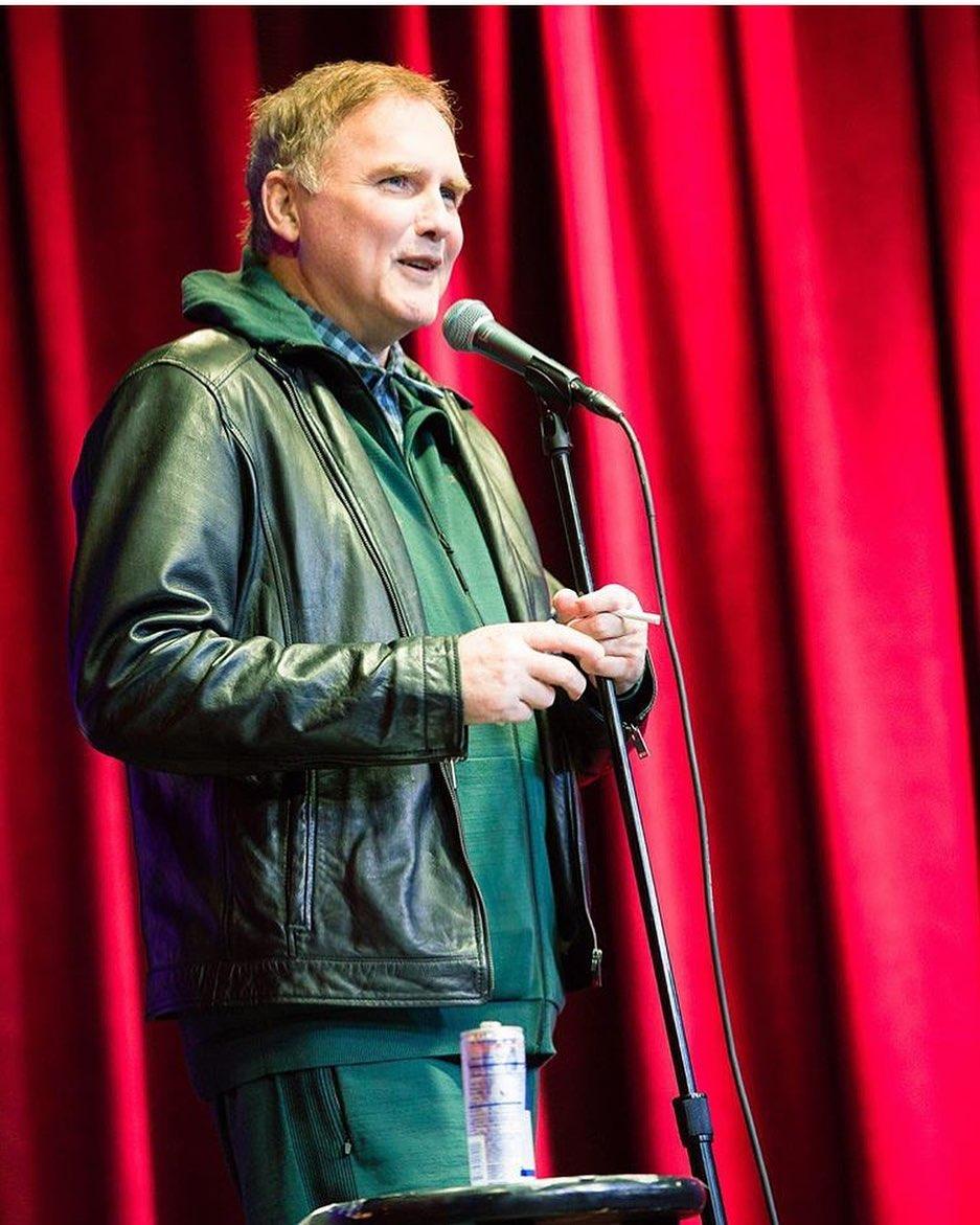 A photo showing Norm MacDonald in a black leather jacket with matching pant, telling jokes on stage.