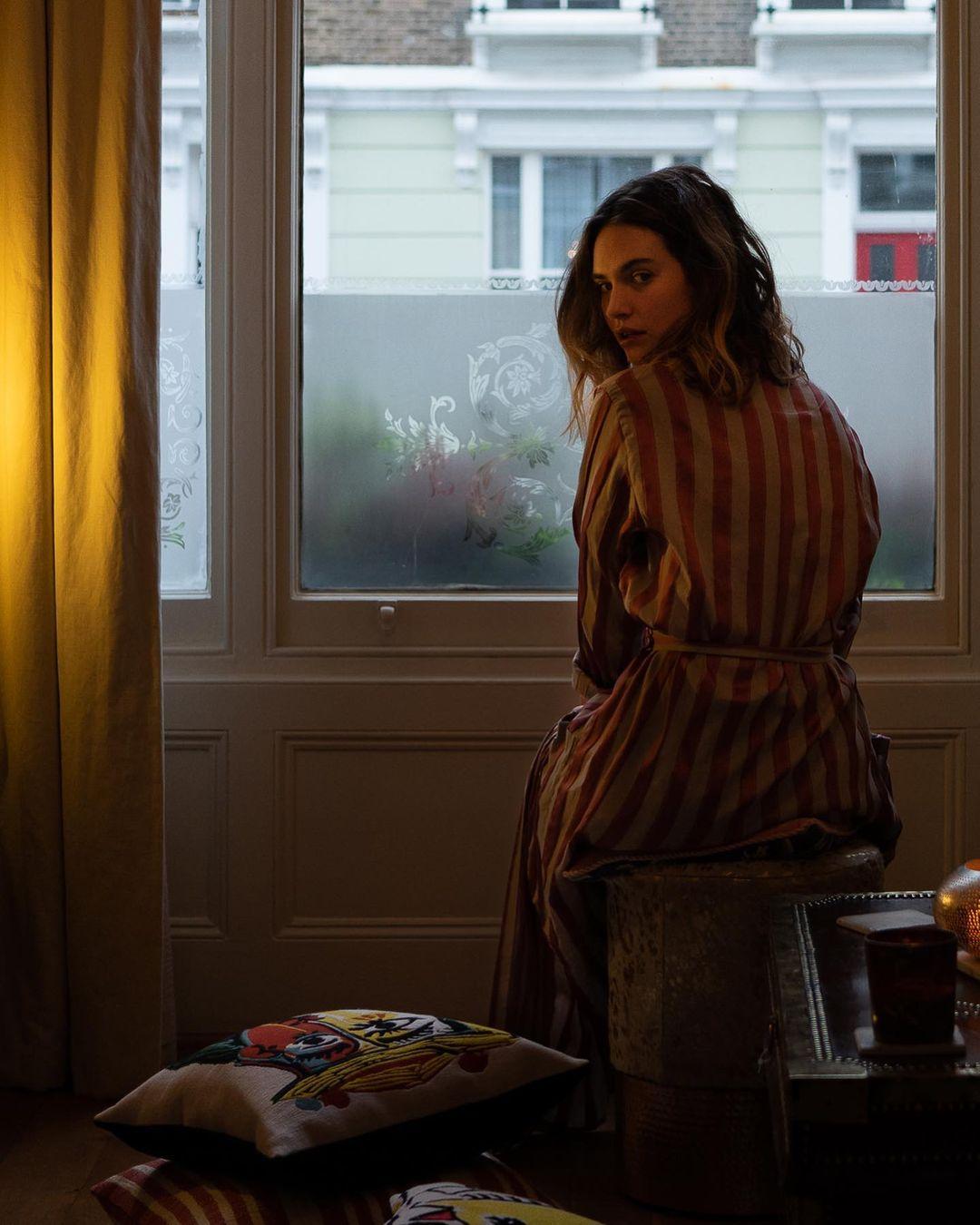 A photo showing Lily James in a brown striped robe, sitting close to the window in her bedroom. 