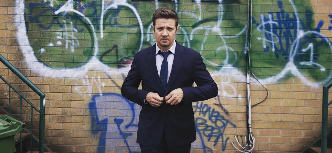 A photo showing Jeremy Renner in a black suit and pant, with a tie to match.