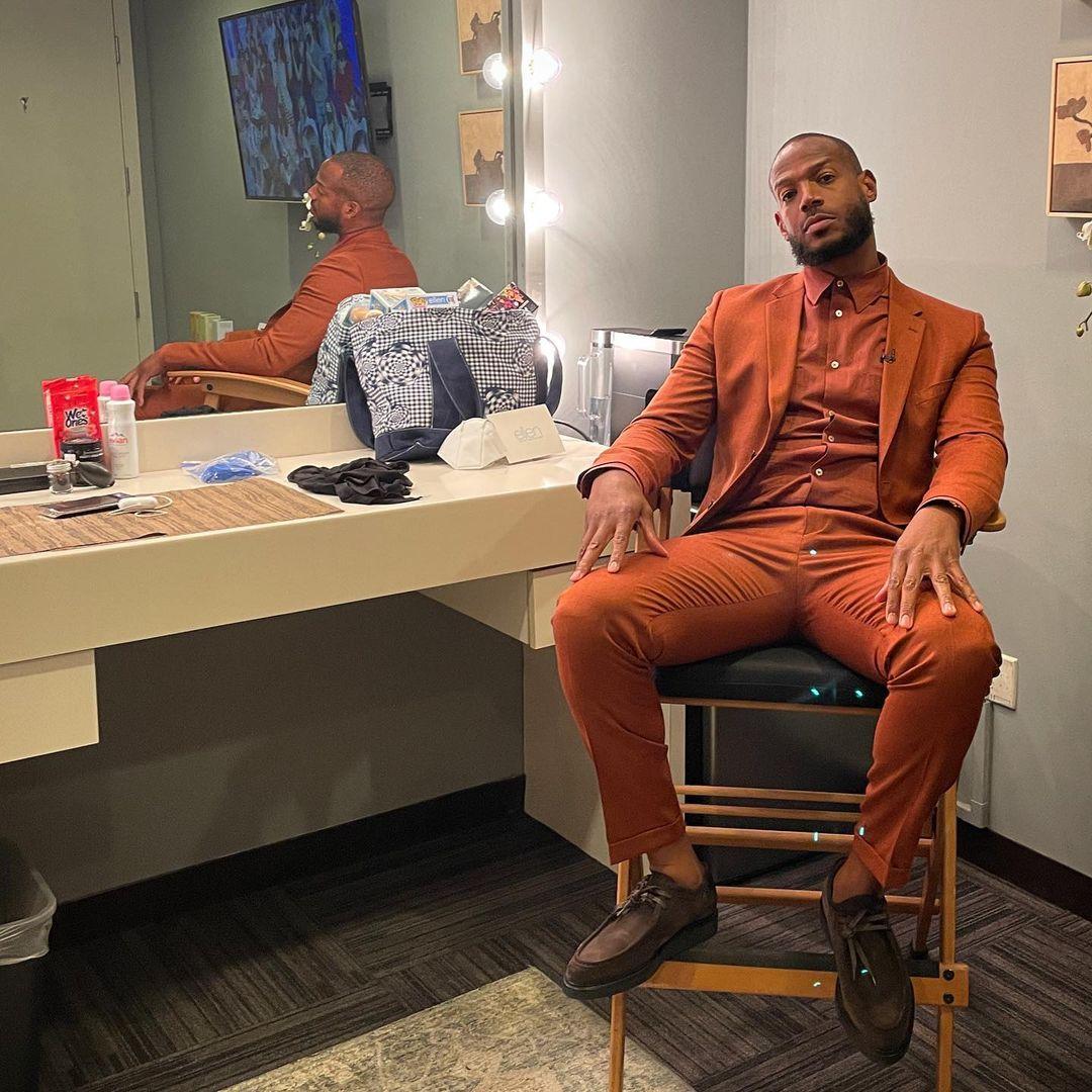 Marlon Wayans looks incredible sitting beside a mirror in a brown suit and pant.