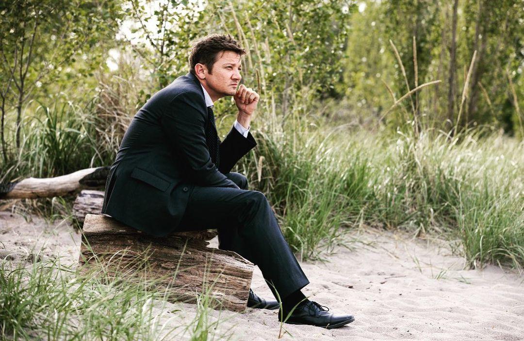 A photo showing Jeremy Renner in a black suit and pant, sitting on a rock.