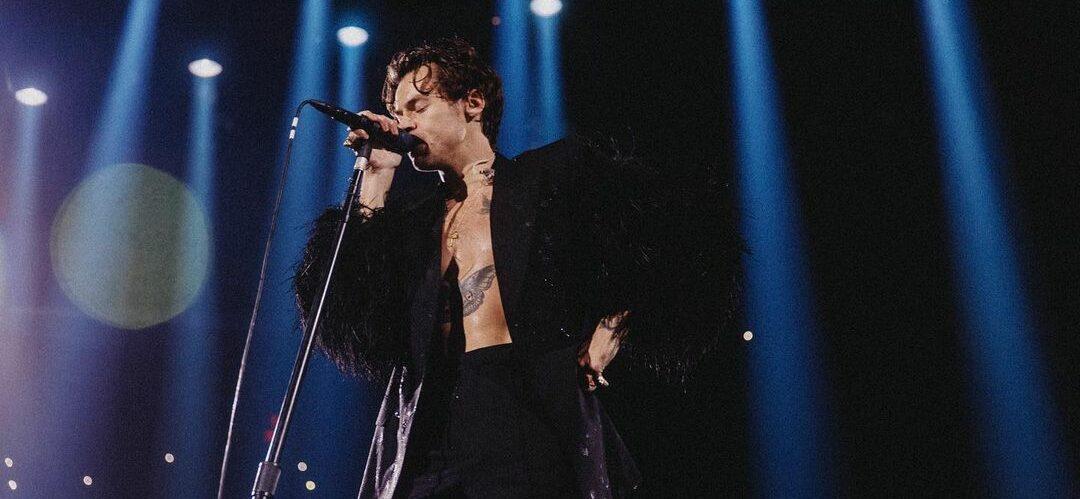 A photo showing Harry Styles in a black jacket and pant, singing on stage.