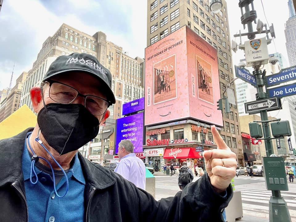 A photo of Ron Howard in a blue T-shirt and jacket, with a facemask, outdoors.