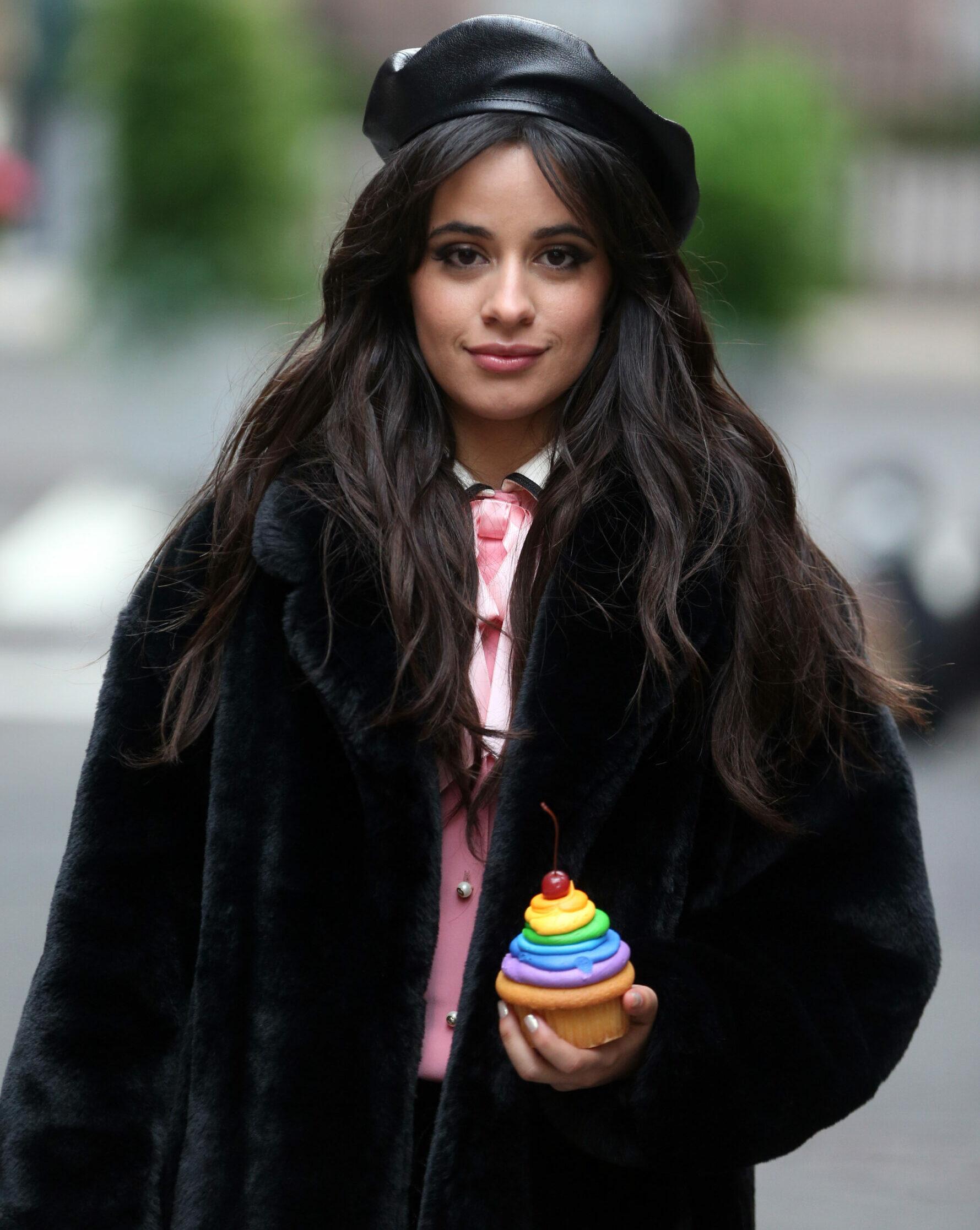 A photo showing Camila Cabello in a black fur jacket and hat to match, holding a rainbow cup-cake.