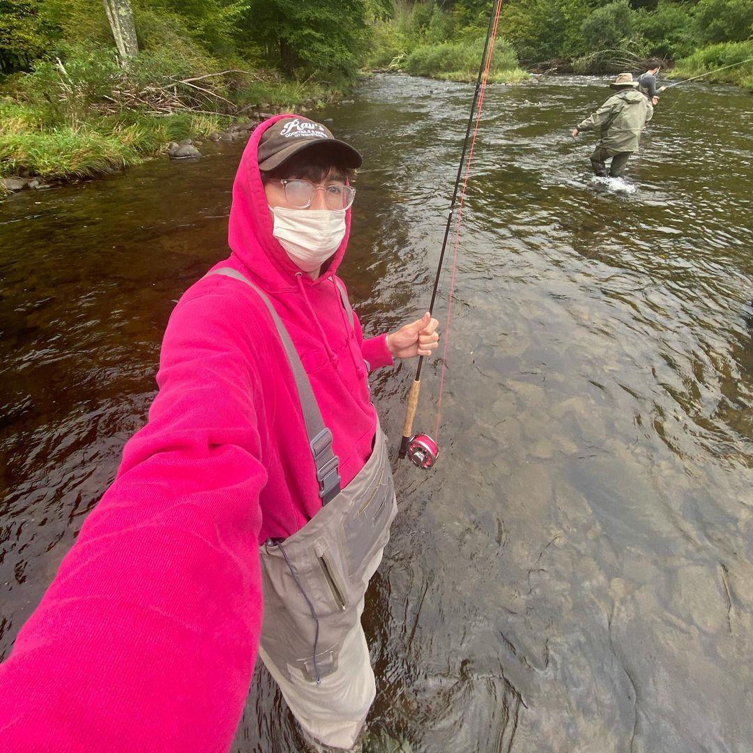 A photo showing Nicholas Braun in a pink hoodie and suspenders trying to fish in a stream.