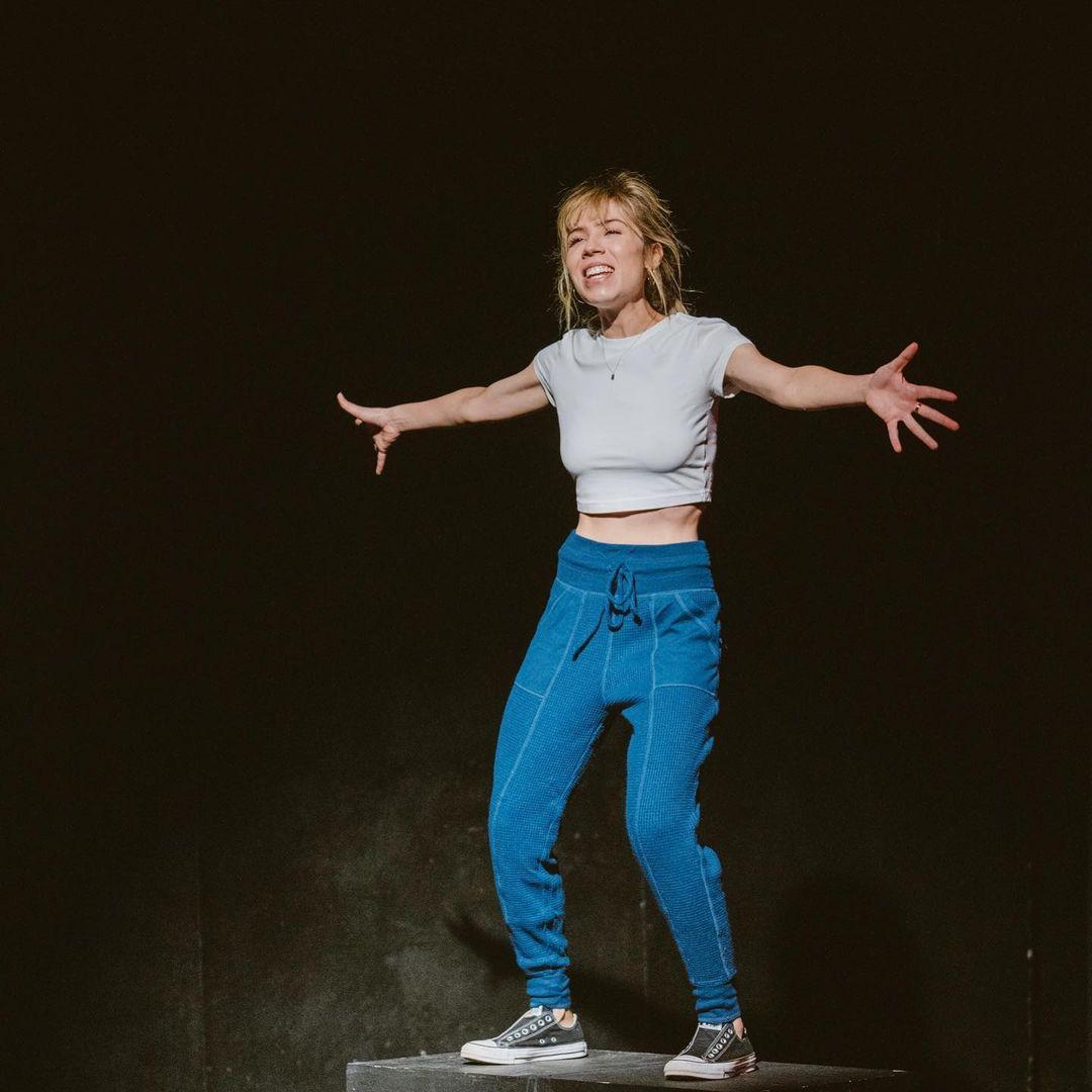A photo showing Jennette McCurdy in a white crop-top and denim pant, with her arms spread apart.