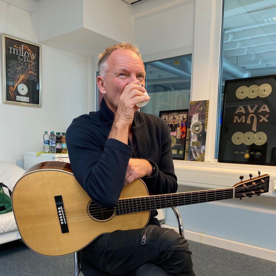 A photo showing singer and actor, Sting drinking from a cup while holding a guitar.