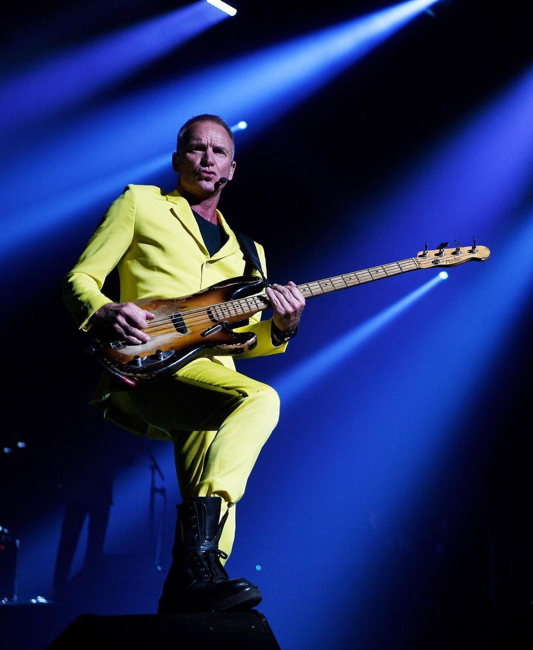 A photo showing singer, Sting, in a yellow suit and pant, with a guitar on stage.