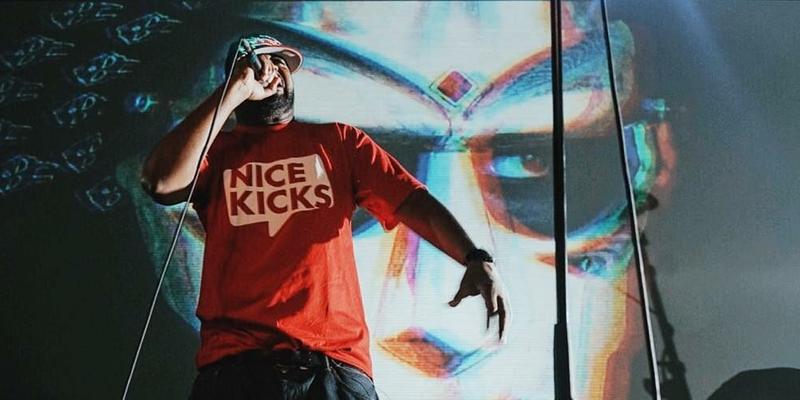 A photo showing MF Doom in a red shirt and black pant performing on stage.