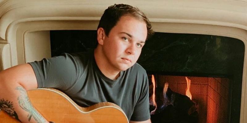 A photo showing Tommy Prine holding a guitar beside a fireplace.