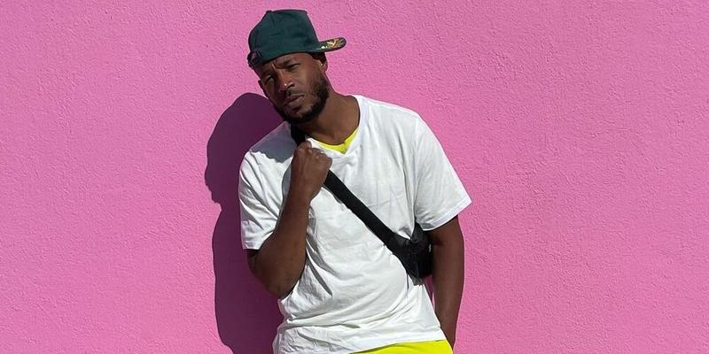 A photo shwoing Marlon Wayans sporting a white shirt and yellow pant, while leaning against a pink color wall.