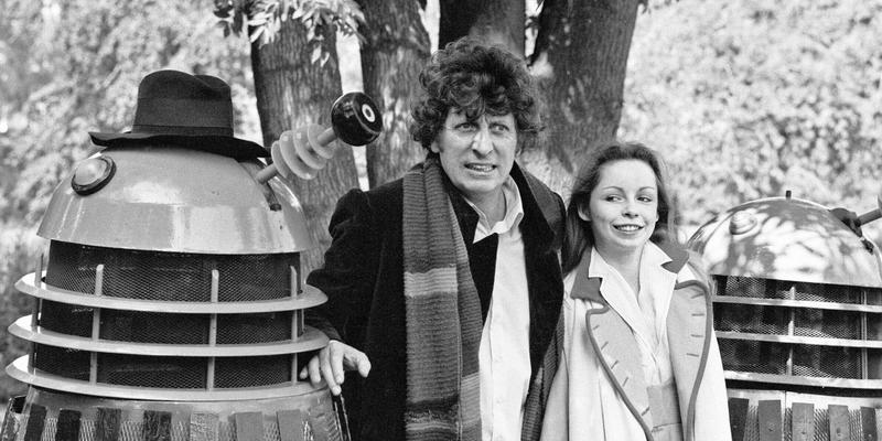 Tom Baker poses as the Fourth Doctor in Doctor Who next to a Dalek and Sarah Baker